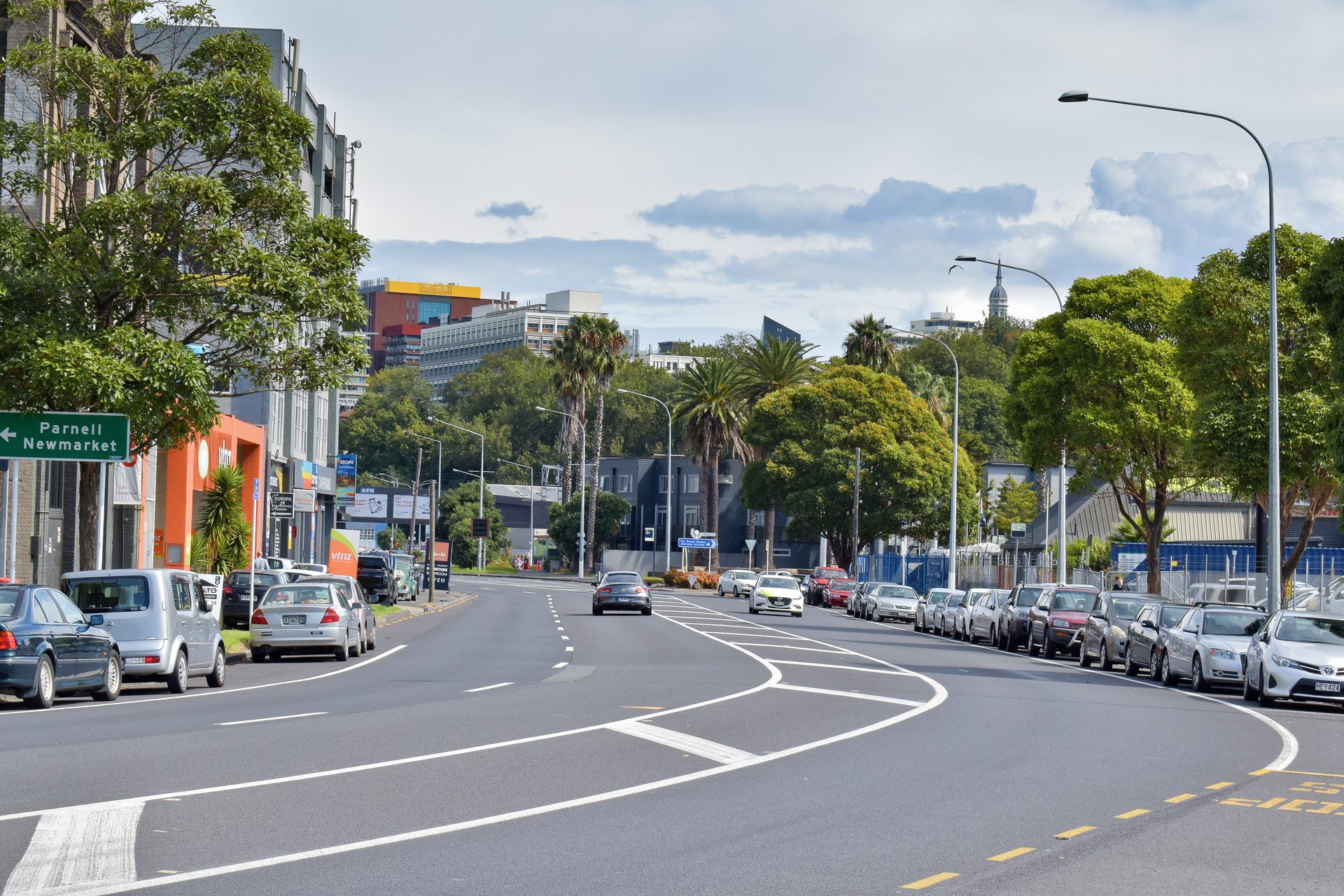 A curving inner Auckland road lined with parked cars and shopfronts, with a green road sign pointing left to Parnell and Newmarket